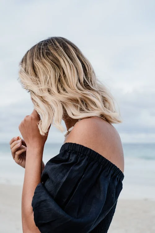 Blonde woman on the beach wearing a black top and adjusting her earrings